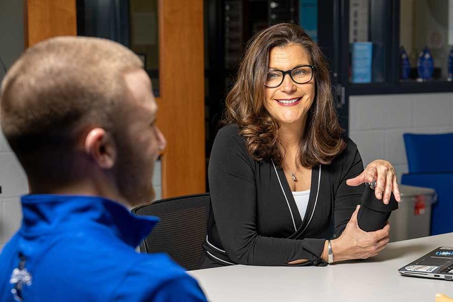 A business professional woman with long dark hair wearing dark glasses sits at a table. Across the table from the woman is a man short hair wearing a blue collared shirt.