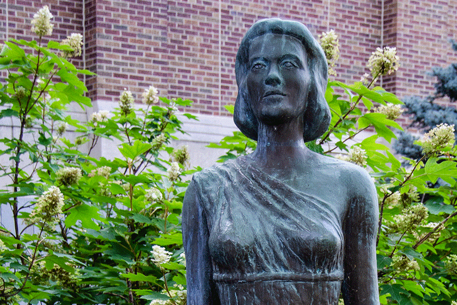 A concrete statue of a woman wearing an off-the-shoulder toga. The statue depicts shoulder-length hair on the woman. Behind the statue, green plants are visible, and a brick building can be seen in the background.