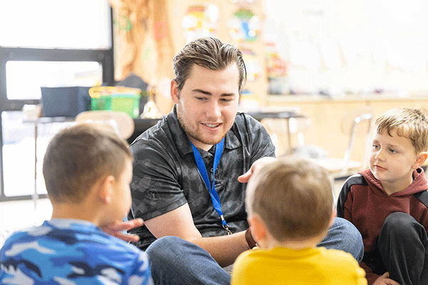 A white, male Indiana State student-teacher, with brown hair and wearing a gray camo t-shirt and blue lanyard, sits and talks with two young boys in a classroom. He is smiling.