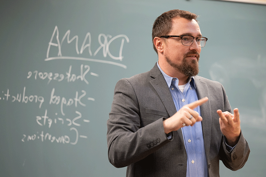 A white male professor with short, dark brown hair and glasses stands at the front of a classroom with the words Drama, Shakespeare, Staged Production, Scripts, and Conventions written on the chalkboard  behind him. He wears a grey suit jacket with a blue open-collared shirt, and his left hand is pointing to his right hand, on which three fingers are held up as if counting.