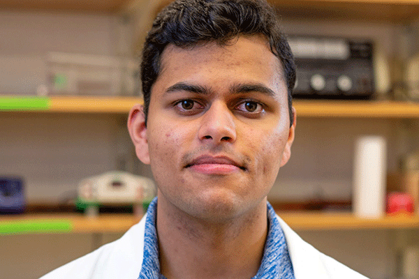 A close-up picture of a male student of Indiana heritage staring directly into the camera with a soft smile. He is wearing a blue shirt and white lab coat. 