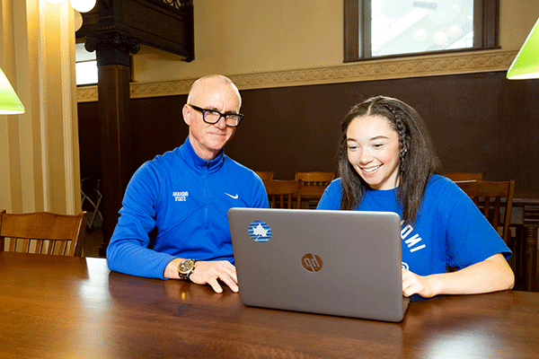 A student in a blue shirt sitting at a laptop and smiling while a male with white hair and a blue shirt looks on. 