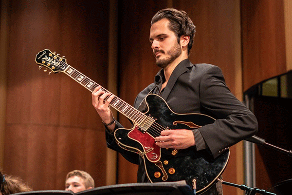 A male with a black shirt and dark hear with beard and a mustache plays a black guitar.  