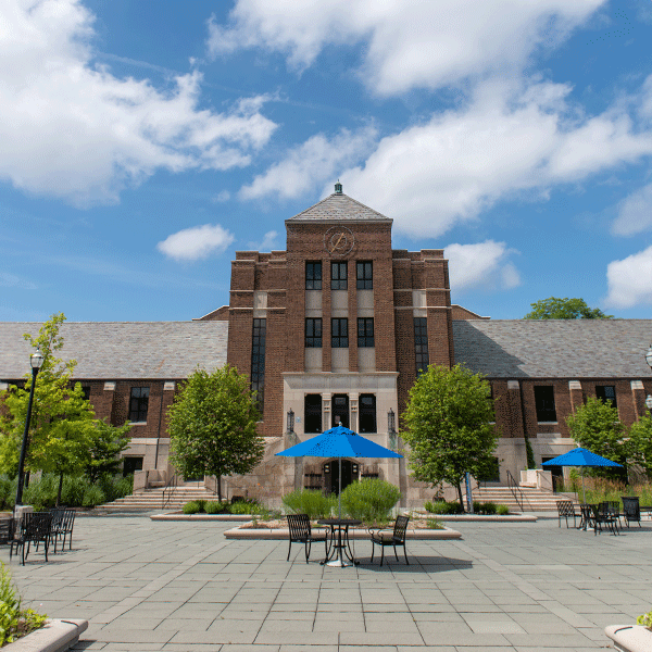 The exterior of a multi story brick building with a patio in front, and la large staircase on the left and right side leading up to the doorway. The sky is blue with white fluffy clouds, and there is a clock tower near the top of the building.