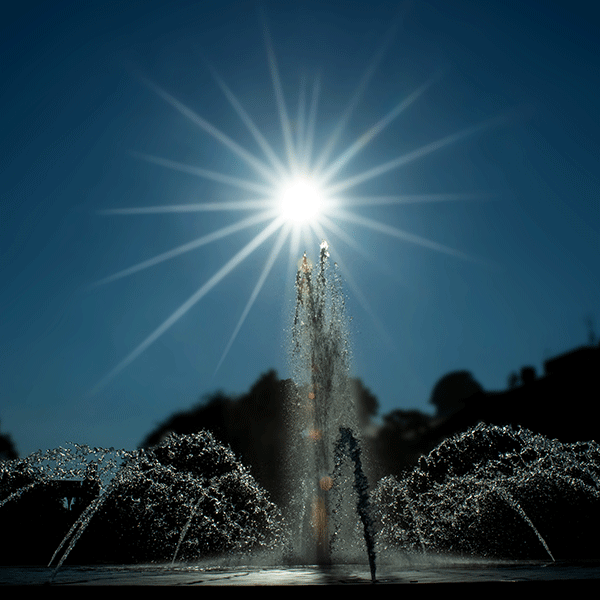 Darkly lit photo of a fountain with a center spout and smaller spouts around the center in a circular pattern with the sun behind with sunbeams extending in all directions from the center of the sun.