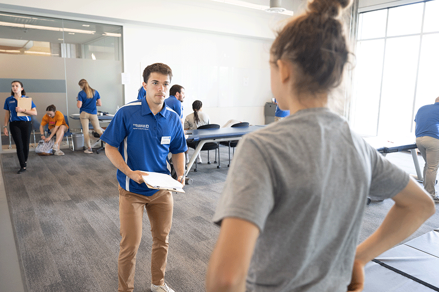 A male student with a blue polo shirt standing in the middle of a large examing room and talking to a clent with her back turned to the camera. 