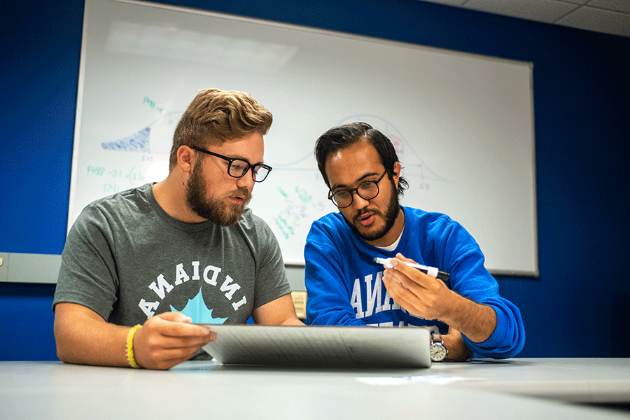 Two men sitting together in a classroom look like they are solving a problem on the white erase board in front of them. The man on the left has an olive complexion, black hair, beard, and moustache, and wears glasses. He is wearing an Indiana State sweatshirt with the sleeves rolled up and holds a black dry-erase marker in his hand. 