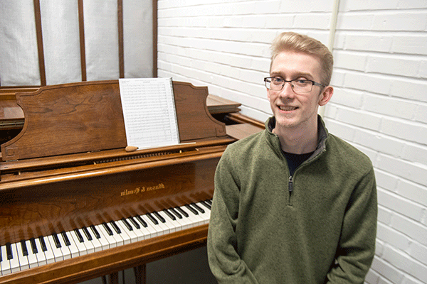 A white male student with blond hair and glasses sits on a piano bench in front of a brown piano. A book of sheet music sits atop the piano. There is a white brick wall in the background.