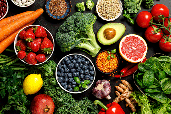 A colorful assortment of fruits and vegetables on a table. Visible items include: red tomatoes on the vine; an avocado cut in half; a pink grapefruit cut in half; green, leafy spinach; lettuce; brown parsnips; bowls of blueberries and strawberries; broccoli; a dish of white beans; a dish of seeds; lemons; a red pepper; a purple and white mushroom; a pomegranate; and some carrots, among other foods.