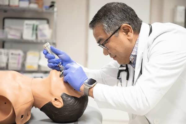 Middle aged male with white coat using a medical instrument to look down the throat of a medical practice mannequin.