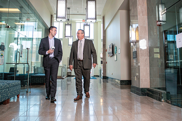  A white male professor in professional business attire walks down a brightly lit hallway with a white male student in professional business attire holding a coffee cup.