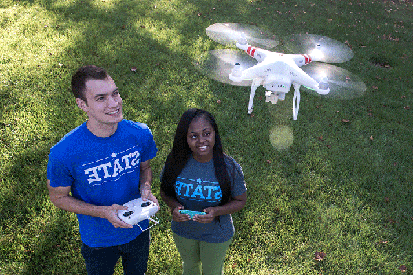 Two students are standing outside on a grassy lawn. On the left is a Black female student with long black hair. She wears green pants, red shoes, and a blue T-shirt with “STATE” in light blue lettering on the front. A blue smartphone is visible in her hands. On the right is a white male student with short brown hair. He wears denim jeans, black shoes, and a blue T-shirt with “STATE” in white lettering on the front. He holds a white drone remote controller while a white drone is flying above the students.