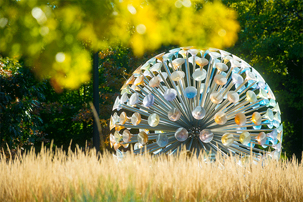 Photo of a large, circular structure made up of mall colorful disks suspended by metal posts installed in a round core, with brown native grass in the foreground and green foliage from a tree in the top right corner. 