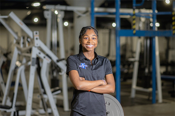 Black female wearing black polo with the Sycamores logo standing with arms crossed and smiling at the camera with exercise equipment in the background. 