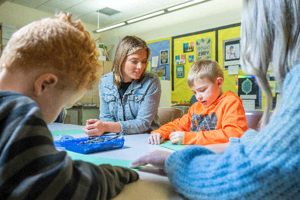 A blonde teaching student with short hair and wearing a blue jean jacket is seated at a table in a classroom. She watches as a young blond boy in an orange sweatshirt seated next to her creates a drawing on green construction paper. In the foreground, another young boy with short red hair works on a drawing, while another female teaching student with long blonde hair and wearing a blue sweater holds the paper in place for him. 