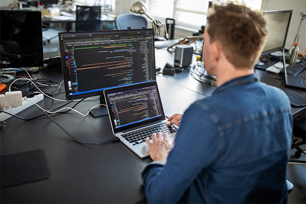 Male professional sitting with back to the camera and working at a laptop with additional computer monitor behind at his office desk in an office setting. 