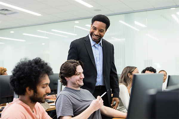 Smiling mature adult male university professor checks in with two male students in a computer lab setting.