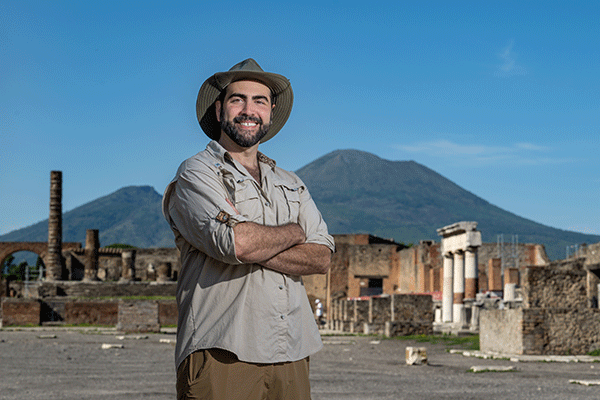 A male professor stands with arms crossed facing the camera in an open area within the ruins of Pompeii with Mt. Vesuvius visible in the background. 