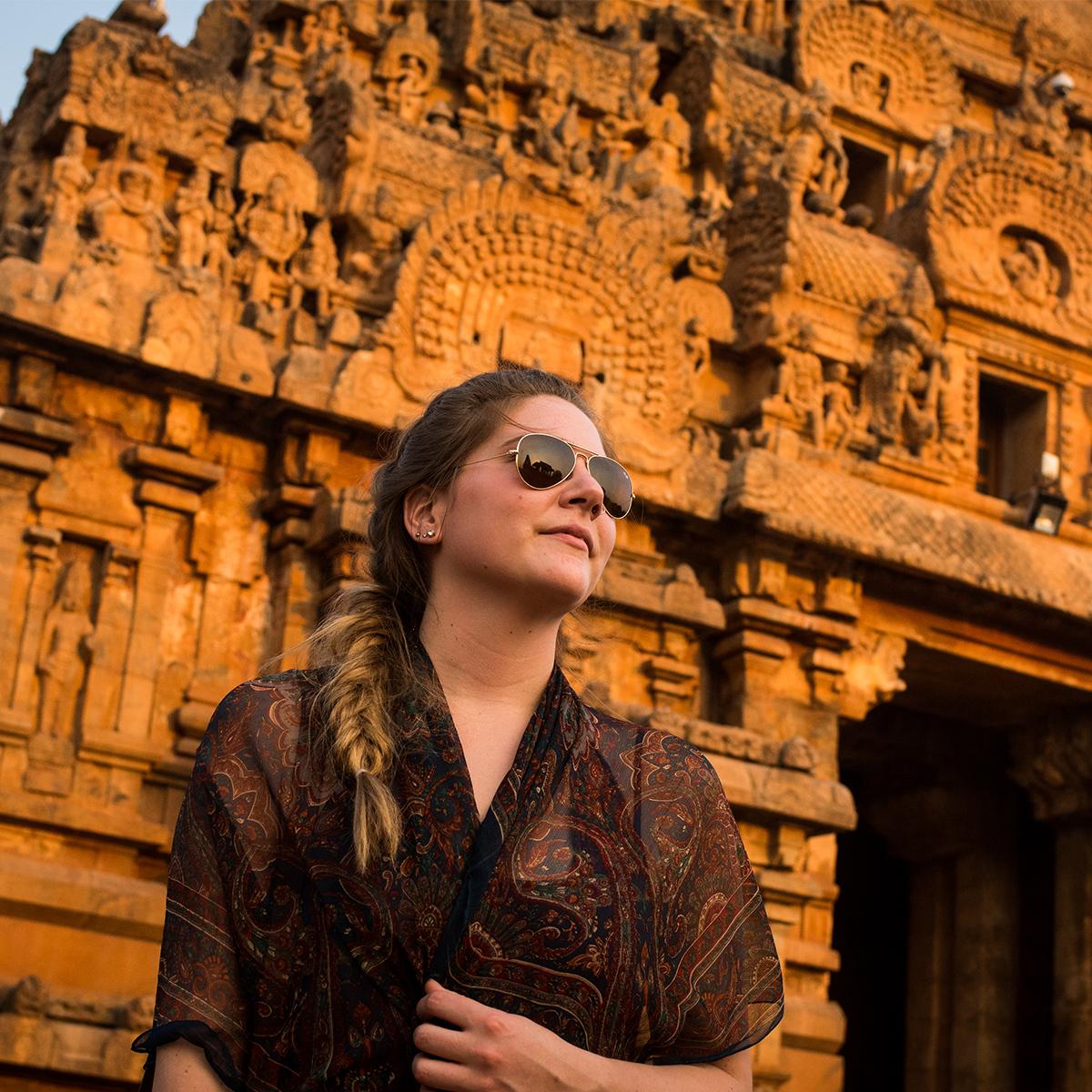 A young woman in aviator sunglasses looks at the setting sun with an ancient temple behind her illuminated orange by the setting sun.