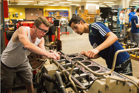 What looks like a a radiator or air compressor – grey with round and square uniform openings in the top – and with assorted green and black valves, hoses, and attachments – sits in a large industrial workspace with yellow walls. Two male students wearing safety goggles lean over it with their hands on it, inspecting something on the far end. The student on the left has short black hair and some facial hair and wears safety goggles and a blue-and-white striped shirt.