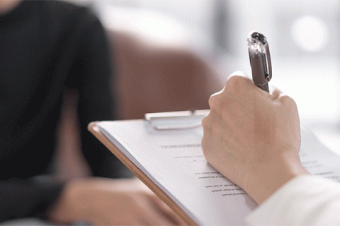 Closeup image of a right hand holding a pen and writing on a form with a clipboard, with a person sitting on a couch out of focus in the background. 