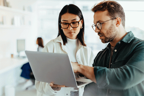 A man with brown hair, a beard and mustache, and glasses, wearing a black shirt, points at a laptop while a woman with straight, shoulder-length, dark brown hair and glasses, wearing a white turtleneck and white jacket, holds the laptop. They are in a bright office setting.  