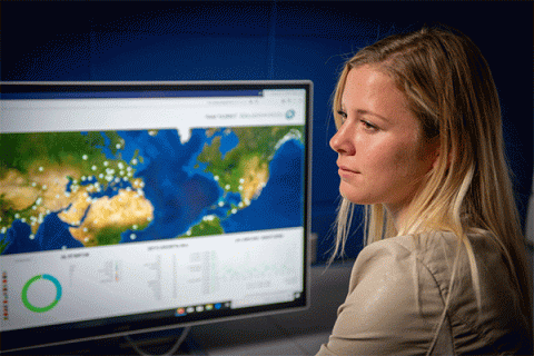 Blonde female student seated in front of a computer monitor and looking to the right with a map showing cybercrime activity on a screen. 