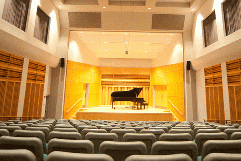 An empty recital hall with a piano in the center of a stage in the background, with rows of seats in the foreground.