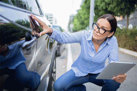 Female middle-aged woman with a tablet in her hand kneeling down and inspecting minor damage on the door of a vehicle. 