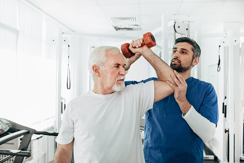 Senior man with white hair lifting a dumbbell, with assistance from an occupational therapist in a medical setting. 