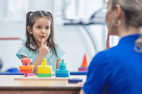 A young child, a girl with brown hair, sits at a table with colorful blocks visible on the table. She wears a light blue T-shirt. She presses her index finger against her chin. Seated opposite her, in the foreground and out of focus, is a woman with blond hair. She wears a blue T-shirt. The background is blurred and an orange cone is visible behind the girl to the left.